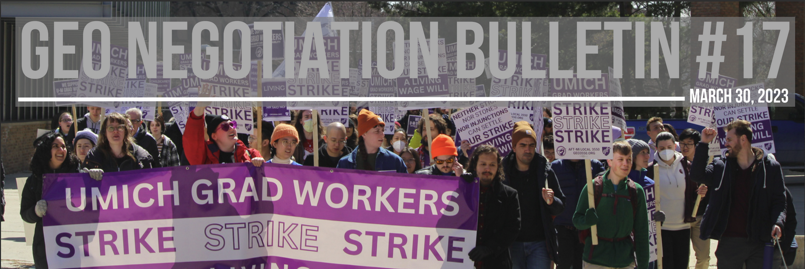 A group of people holding picket signs. A strike banner is prominent at the front