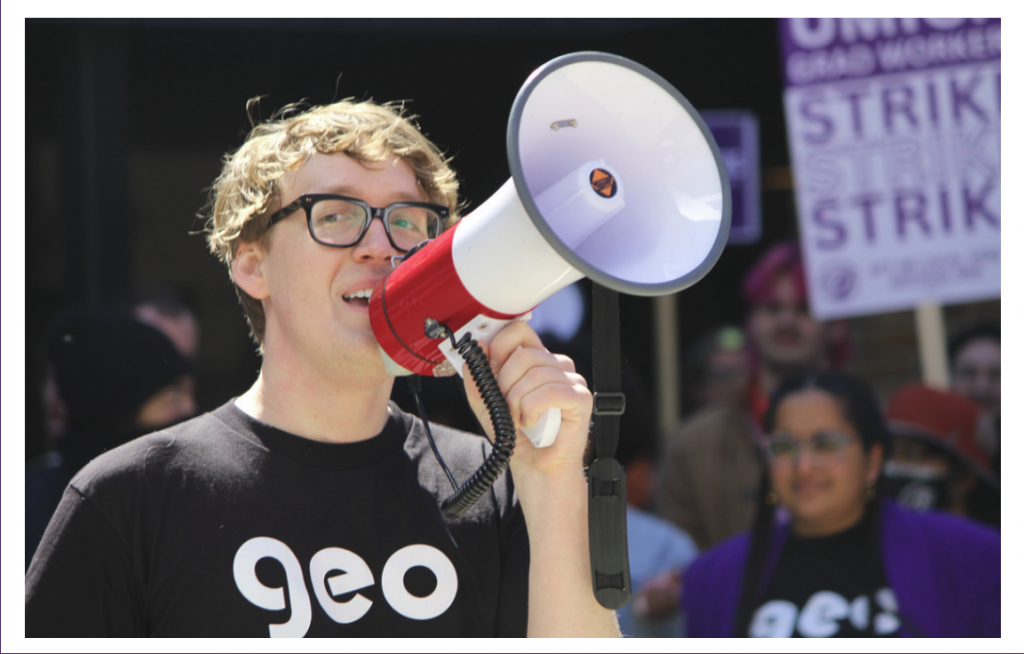 A man with glasses and a GEO shirt speaking into a megaphone. A strike sign is in the background