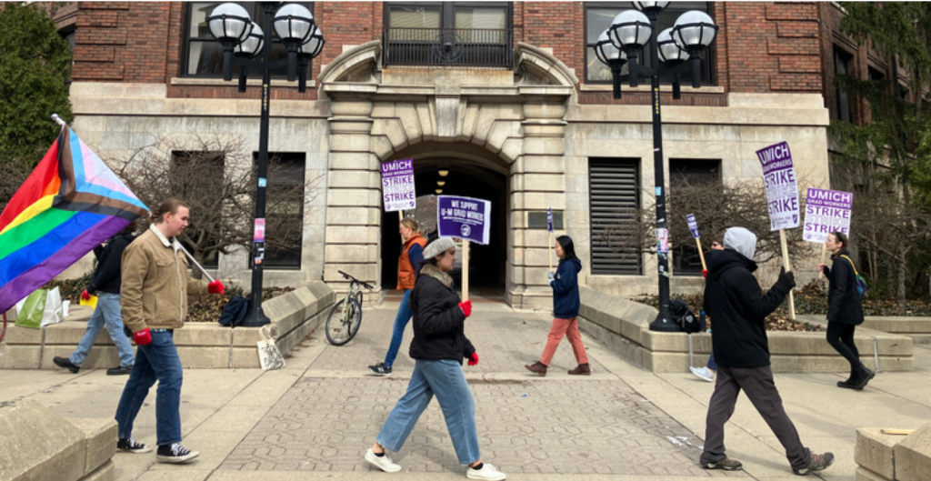 A picket team walking in a large, spaced circle. They are holding strike signs and one is holding a large Pride flag