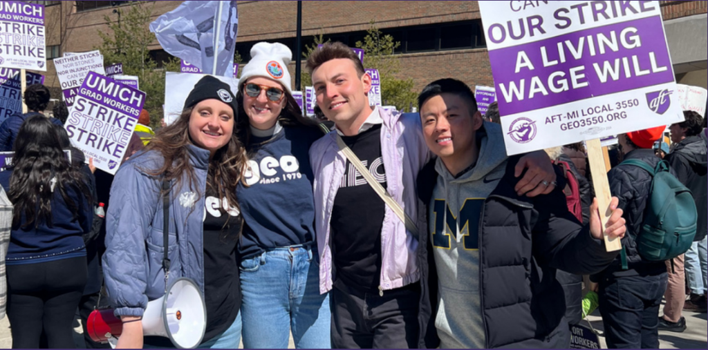 A group of 4 people in posing for a picture with a crowd behind. They have protest signs aloft