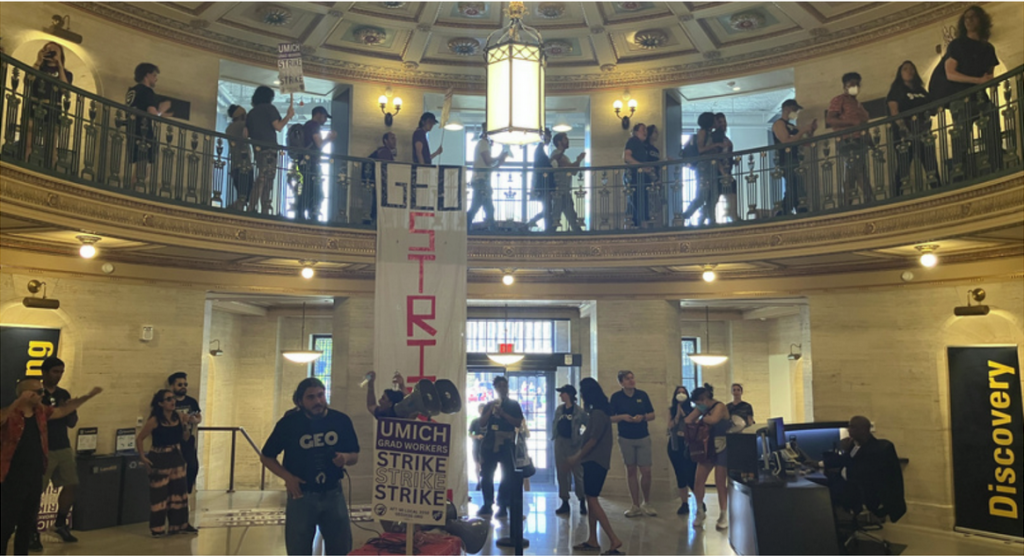 A group of people inside a building on the first and second floor. There is a vertical GEO STRIKE banner hangin off from the second floor