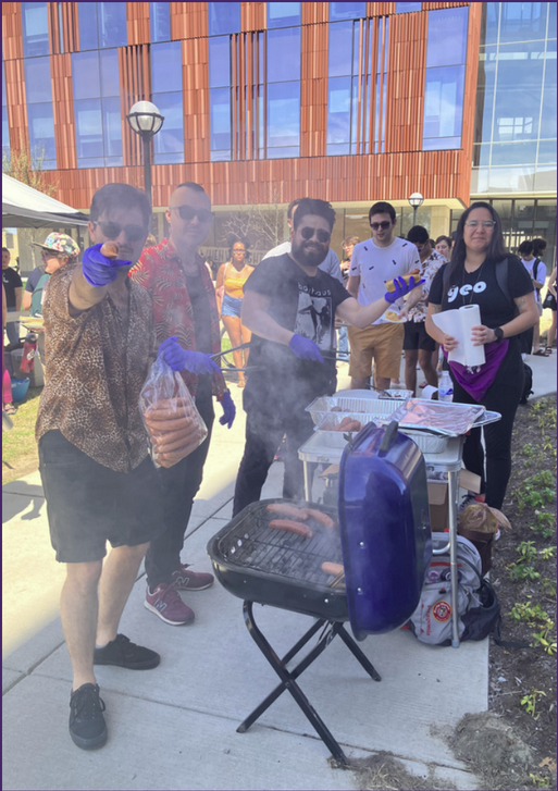 Four people featured in a crowd around a BBQ. One person points tot he camera with a hotdog (in bun)