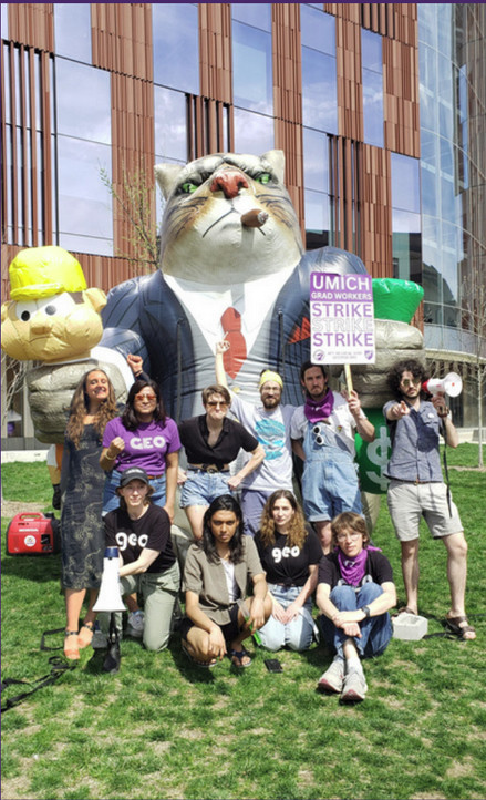 A group of people posing around an inflatable cat. This cat is personified as a boss wearing a suit and cigar choking a construction worker and holding an emerald