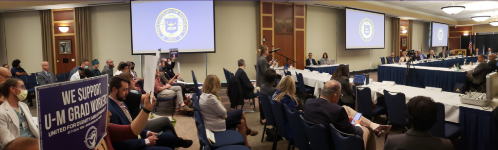 A wide shot of an audience holding "We Support U-M Grad Workers" signs on the left and a board of admin on the right